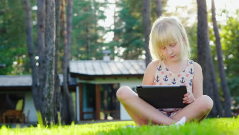 la niña está jugando en la tableta el sol resalta maravillosamente su cabello rubio se sienta en el césped nea