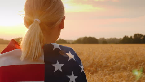 a woman with a usa flag on her shoulders enjoys a field of wheat at sunset