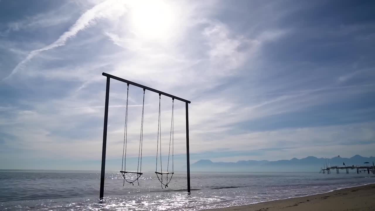 Footage of swings in a beach in water or the sea in Portugal while the water or the sea is moving beneath them and nights and evening sky is visible with clouds in 4K resolution. A pier in the back.