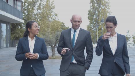 smiling confident business colleagues walking at office building, drinking coffee and talking together