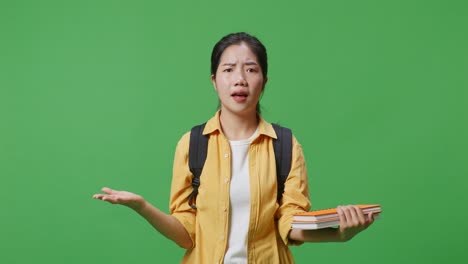 wondering asian woman student with a backpack and some books standing doubtfully saying why in the green screen background studio