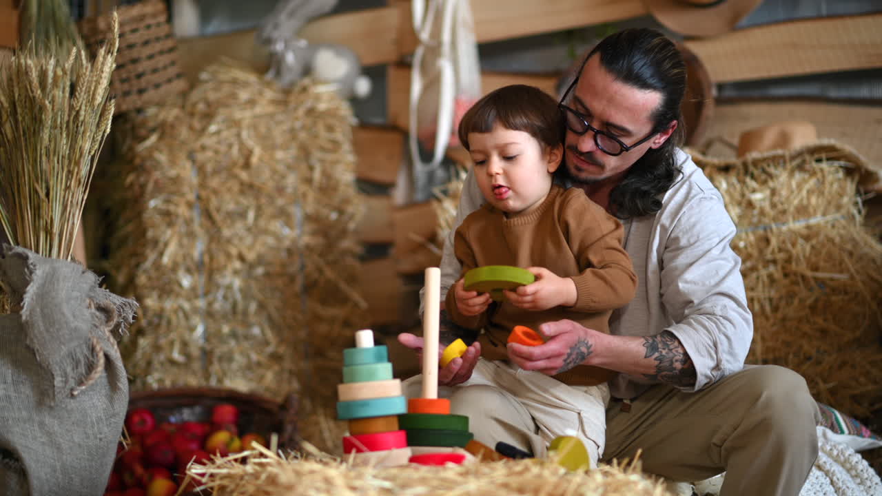 Father playing with his son with colourful, ecological wooden toys in a barn, near square hay bales