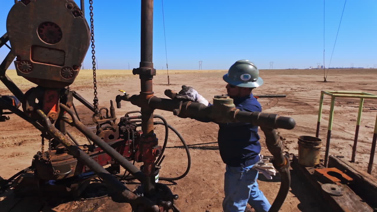 Two men wearing protective helmets work at the oil production site. Worker uses a hummer to unscrew the nut at the metal rusty pipe.