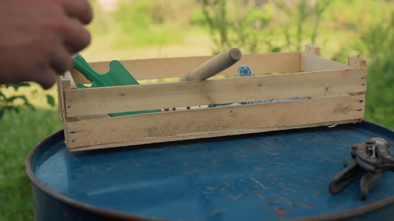 Il contadino ripone la forca da giardinaggio in una cassa di legno su un fusto metallico blu, la mano che fa scorrere il manico tra le doghe, guanti e cesoie nelle vicinanze, allestimento da giardino all'aperto in una calda luce diurna