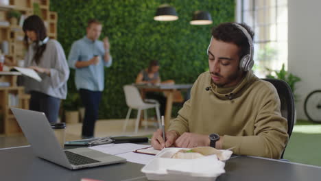 young-arab-businessman-student-brainstorming-writing-notes-enjoying-study-using-laptop-researching-online-listening-to-music-in-busy-office-workplace