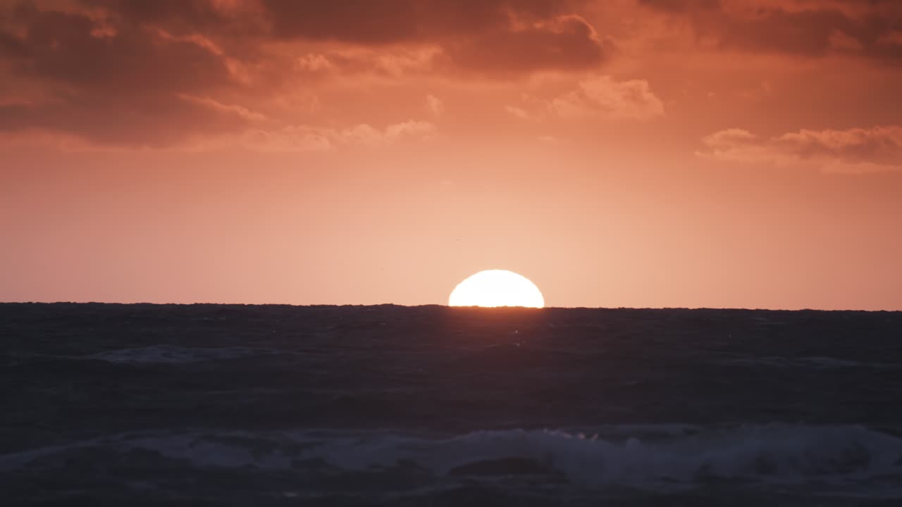 Gentle waves move rhythmically under a glowing sunset, illuminating the sea with hues of orange and blue.