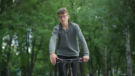 young boy in grey top and black trousers riding bicycle outdoors in park, he stands while holding the bike handlebars, focusing ahead, the lush green trees provide a natural background