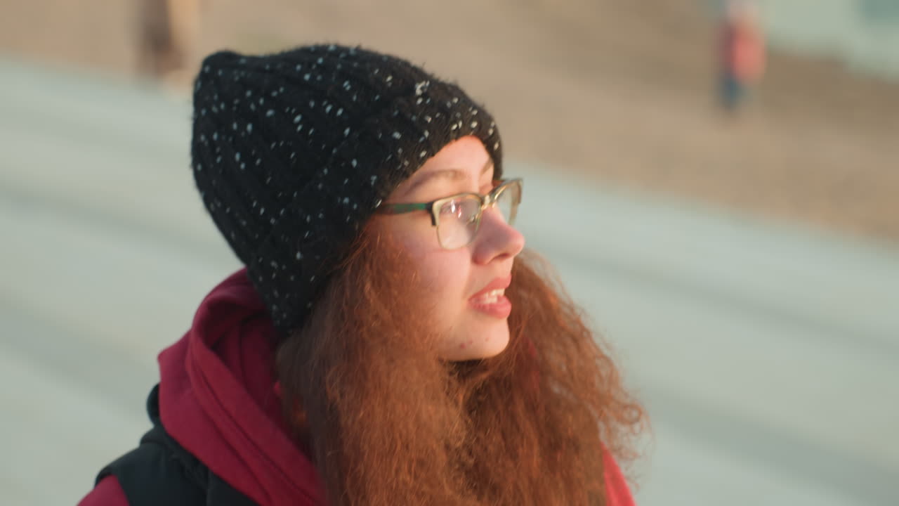 Donna sorridente che si sistema i capelli in spiaggia, con berretto e occhiali, ricci ramati e felpa rossa con cappuccio, momento di vita autentico di connessione e fiducia, relax informale, luce solare soffusa e oceano calmo.
