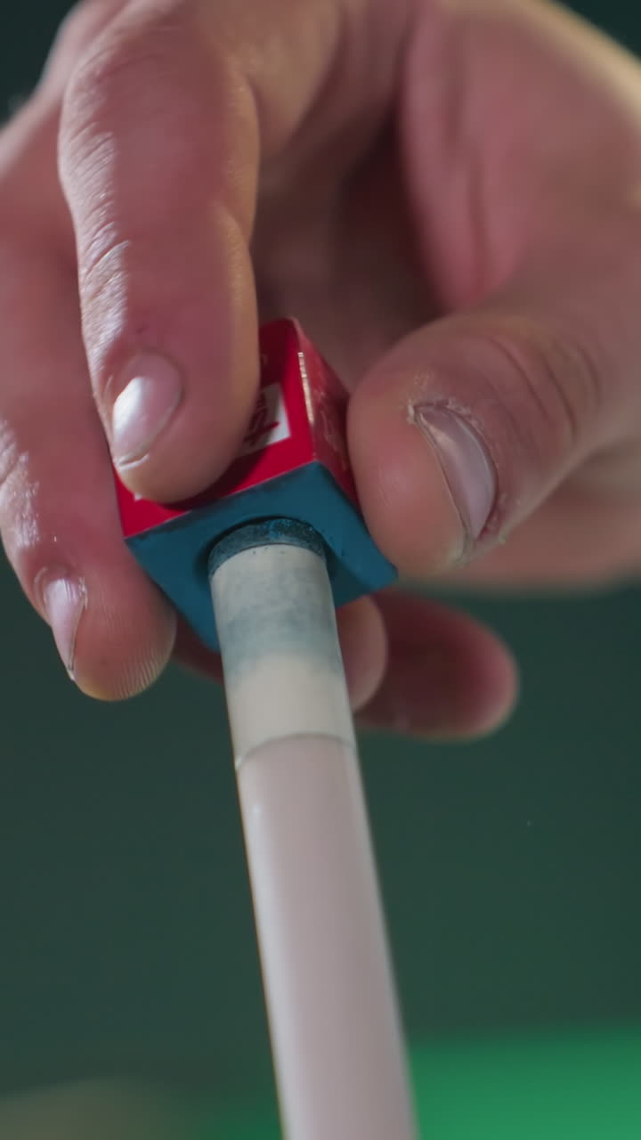 el fondo borroso destaca el primer plano de la mano en camisa blanca aplicando cuidadosamente la tiza al palo de señal antes del juego de billar. la tiza roja y azul contrasta con el agarre enfocado, mostrando la preparación