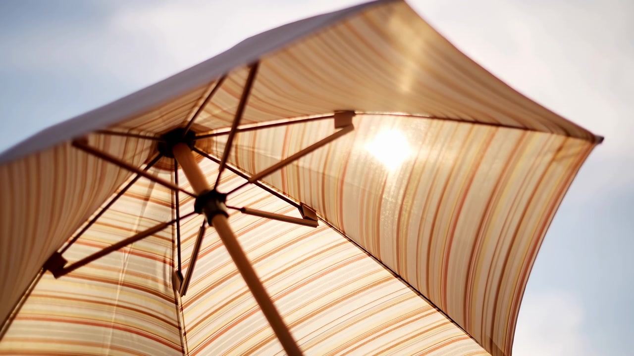 Parasol de plage rayé ouvert vu en contre-plongée, le dessous de la toile translucide brillant sous la lumière chaude du soleil, avec mât central, baleines, connecteurs, texture peinte et fort contre-jour du ciel