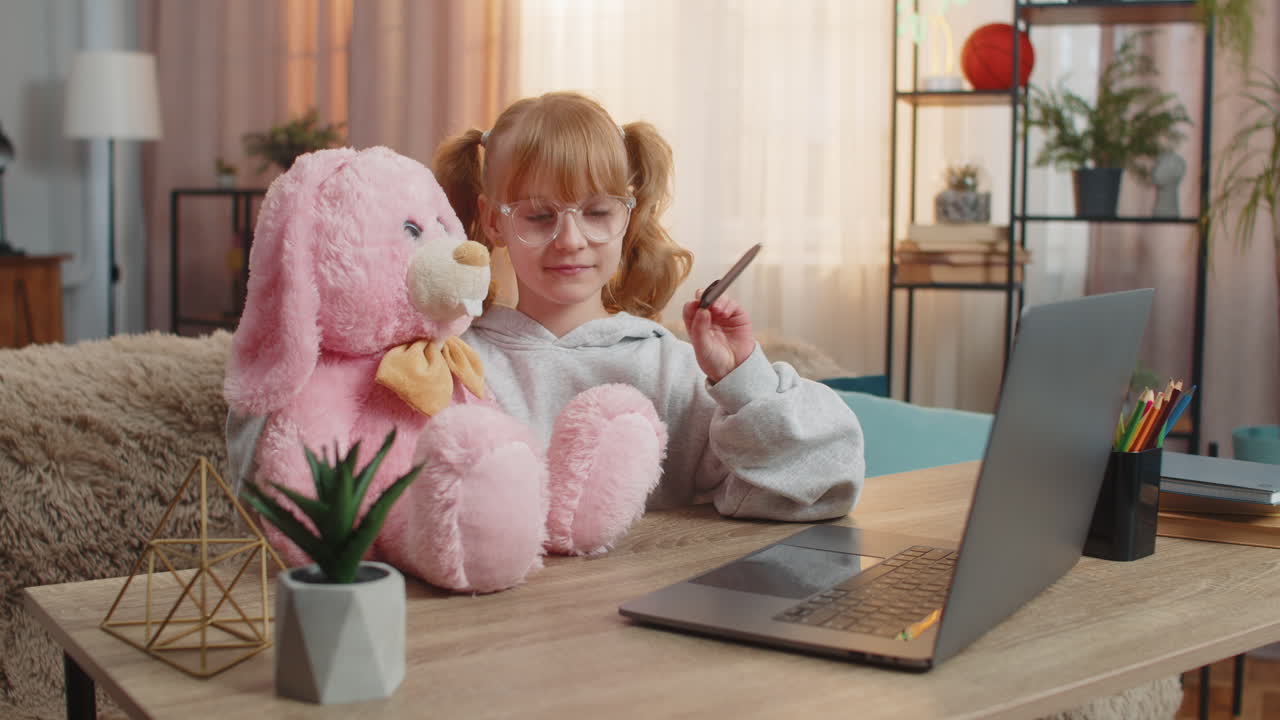 Jovem estudante senta-se com um brinquedo de coelho perto de um laptop assistindo a um vídeo educacional explicando o material