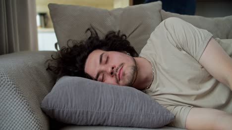 retrato de un hombre moreno cansado y somnoliento con el cabello rizado en una camiseta gris que está cayendo sobre una almohada en el sofá en casa