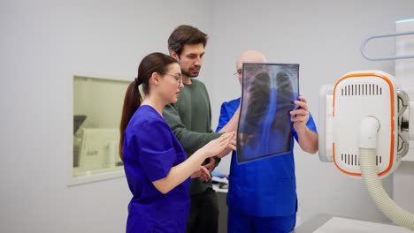 Un-Hombre-Moreno-Con-Barba-Incipiente-Y-Chaqueta-Verde-Se-Comunica-Con-Una-Niña-Y-Un-Médico-Con-Uniforme-Azul-Después-De-Realizar-Una-Fluorografía-Frente-A-Una-Imagen-De-Su-Propio-Pecho-En-Una-Hoja-Transparente-Especial-En-Una-Oficina-De-Una-Clínica-Moderna.