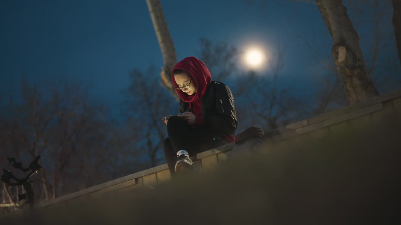 Woman Gazing At Phone Peacefully, Serene Female Figure Observes Phone Amid Moonlit Trees At Night, Quiet Woman Sits On Park Bench Gazing At Her Device Under Distant Moon And Leafless Trees
