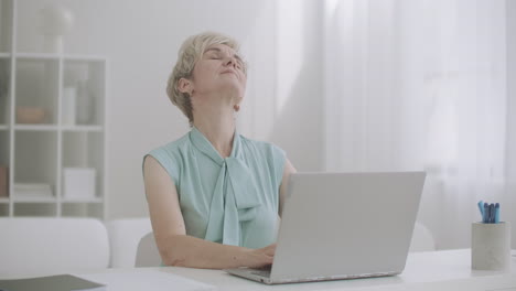 tired aged woman is stretching hands and shoulders during work with laptop sitting at table and typing messages