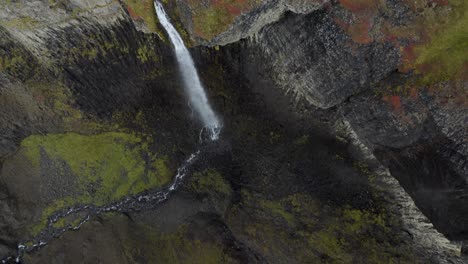 Toma-Aérea-Inclinada-Hacia-Abajo-De-Una-Majestuosa-Cascada-Que-Se-Estrella-Contra-Acantilados-Rocosos-En-El-Valle-De-Islandia