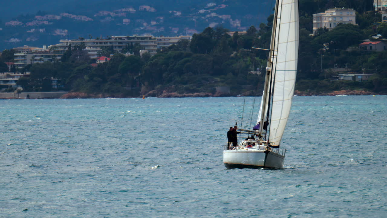 Juan-les-Pins, France - April 3, 2025: Sail boat moving on the sea with the city and the mountain on the background