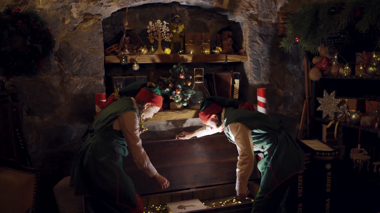 Two elves in a dark room open the big chest which shines inside. Fairy characters taking magic book from the wooden chest on xmas background indoors.