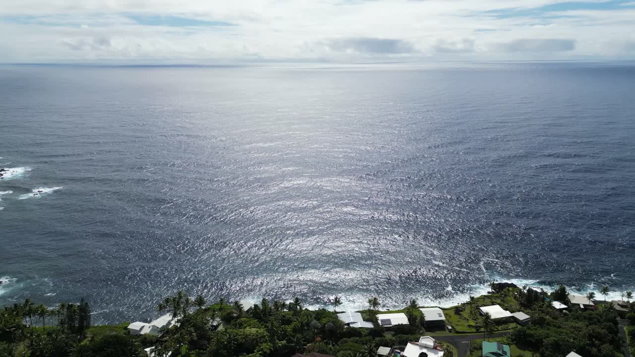 Drone rises above coastal homes on Big Island, revealing wide view of the ocean under bright sky.