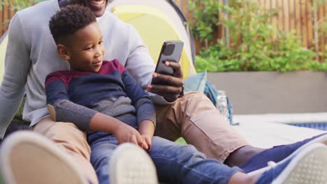 Video-De-Un-Feliz-Padre-E-Hijo-Afroamericanos-Usando-Un-Teléfono-Inteligente-En-El-Jardín