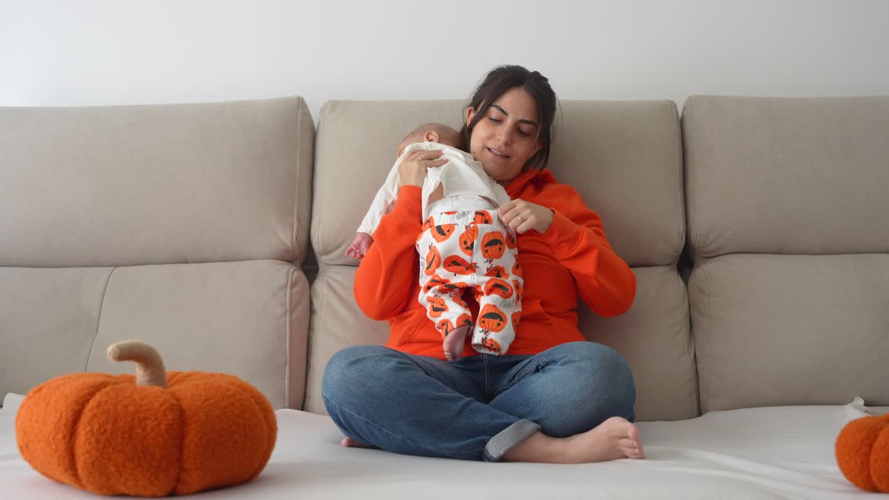 Young mum adjusting the Halloween pants of her 4 months old baby son sitting in the sofa covered with a blanket and pumpkins