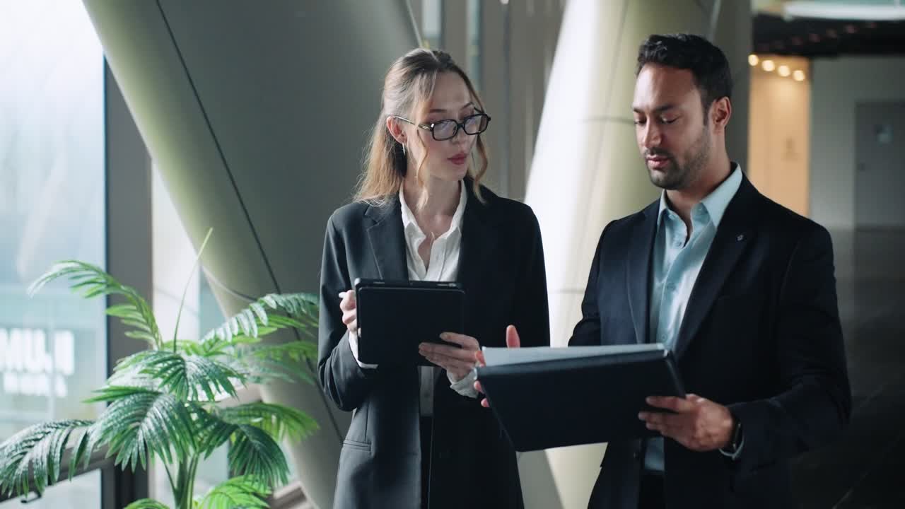 Business coworkers reviewing reports together in office with large windows, discussing and taking notes, medium push in