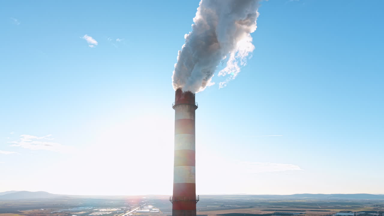 Ciminiera industriale verticale che emette fumo denso contro il cielo blu. Primo piano di una ciminiera industriale a strisce rosse e bianche che rilascia fumo bianco in un cielo blu limpido di giorno