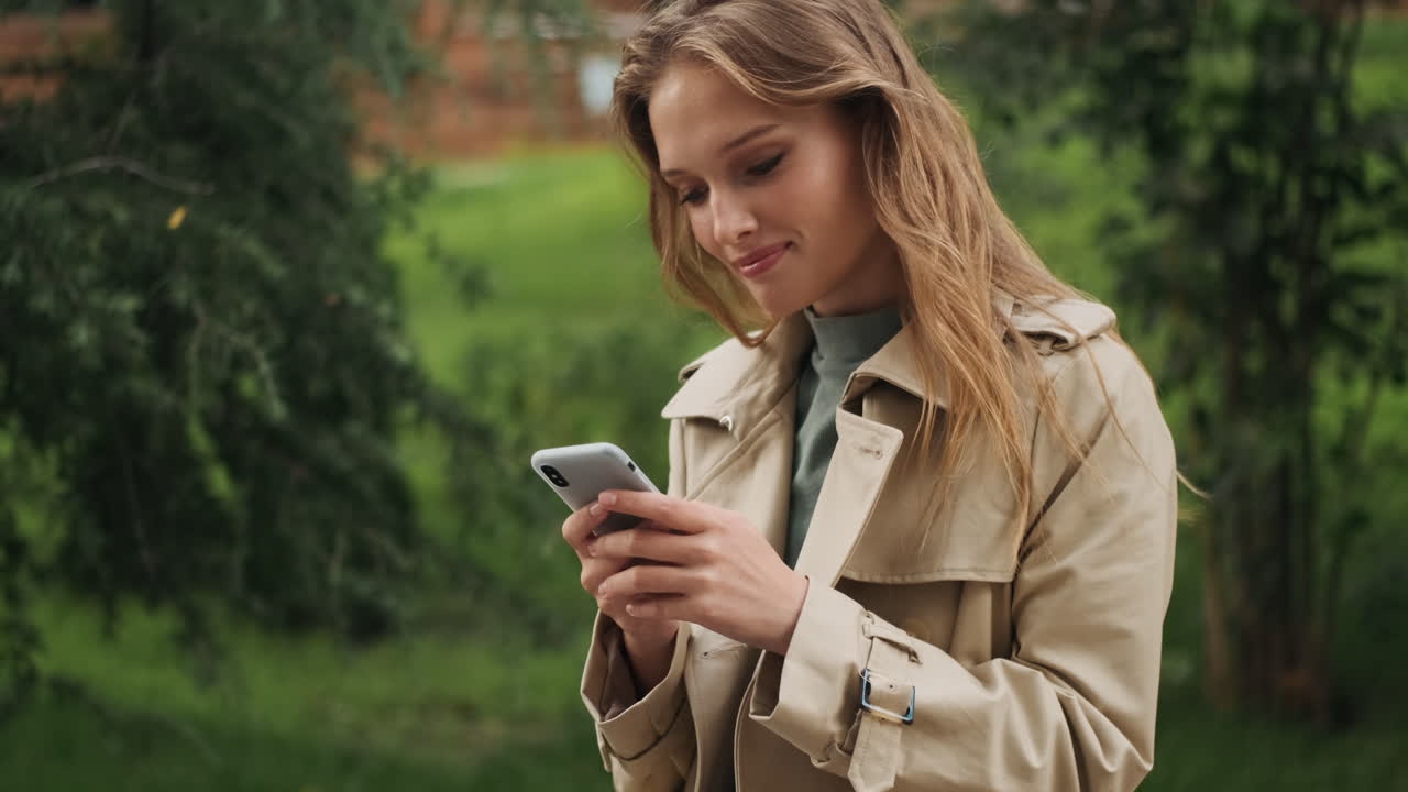 Caucasian female student using smartphone and smiling outdoors.