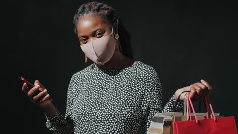 portrait of african american woman in mask with shopping bags and phone