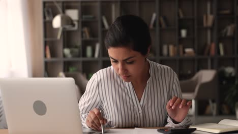 young indian woman sit at desk use calculator calculates expenses