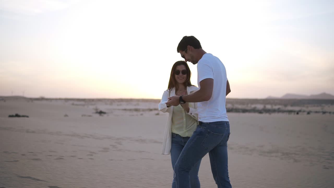 agradable encantadora alegre pareja riendo con camisetas blancas casuales. hombre de cabello oscuro girando con su dama, tomándose de la mano en movimientos. bailando en la naturaleza en el desierto vacío
