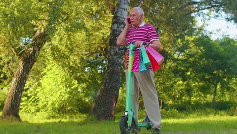 Anciano-Abuelo-Hombre-En-Scooter-Eléctrico-Con-Coloridas-Bolsas-De-Compras-Hablando-Por-Teléfono-Móvil