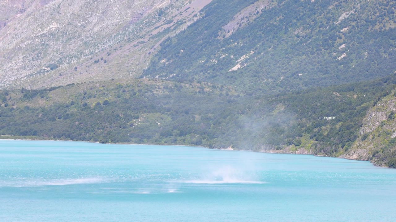 Ampia inquadratura cinematografica di una tromba marina vorticosa che si forma sopra un lago patagonico di un blu vibrante, circondato da aspre montagne andine e foreste in pieno giorno; incontaminato, dinamico, senza persone