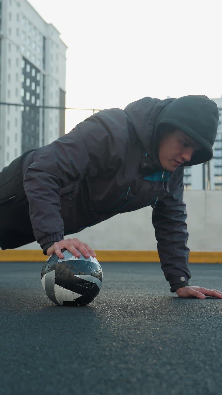 Athlete performing push-ups alternating hand placement on soccer ball demonstrating strength, coordination, and agility in outdoor urban arena with goal post and residential building background