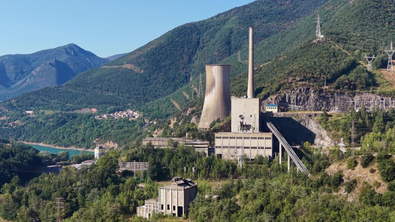 Centrale elettrica a carbone abbandonata a Cercs, Spagna, con una torre di raffreddamento e una ciminiera circondate da verdi valli montane e un bacino idrico turchese