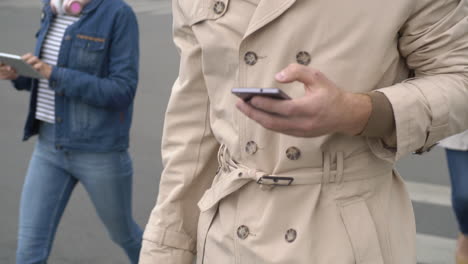 Young-Blond-Man-In-Raincoat-Walking-Down-The-Street-And-Using-Smartphone,-In-The-Background-People-Walking