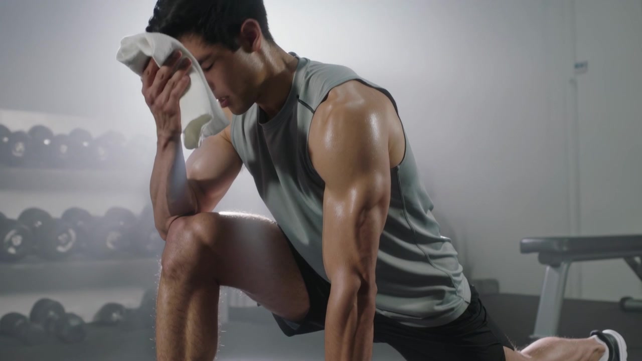 Wiping face after rep, man in sleeveless top holding lunge at gym with white towel recovering