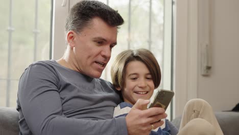 father and son laughing while looking at a smartphone together at home