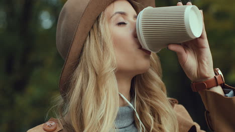 vista de cerca de una joven rubia caucásica con sombrero y auriculares escuchando música en el smartphone y bebiendo café en un hermoso parque de otoño