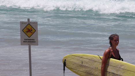 surfer walks by warning sign towards the ocean waves.