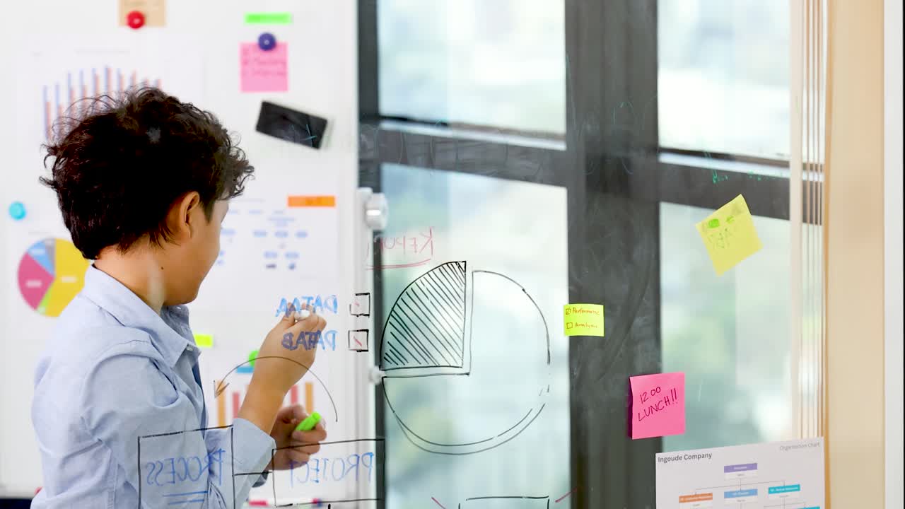 A young boy draws business diagrams on a glass wall in a bright modern office