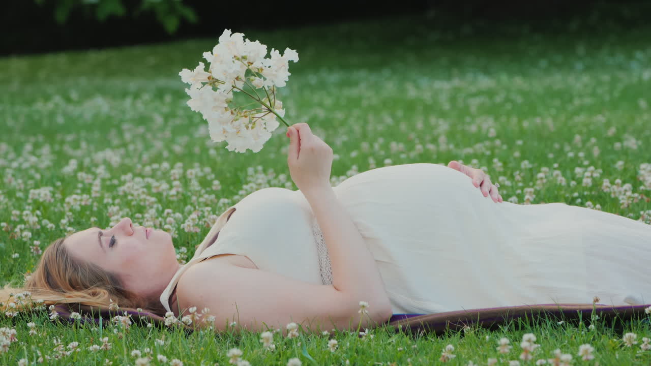 귀여운 임신한 여자는 잔디밭에 누워 그녀의 손에 꽃을 꿈
