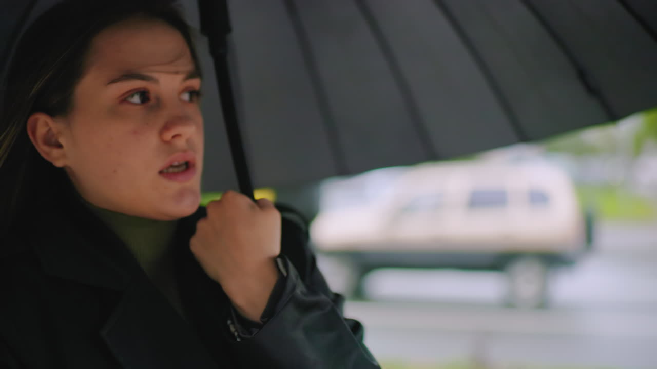 Close up of female in black coat standing under umbrella with hand on city street with blurred white van and trees in background during cloudy day,outdoor lifestyle