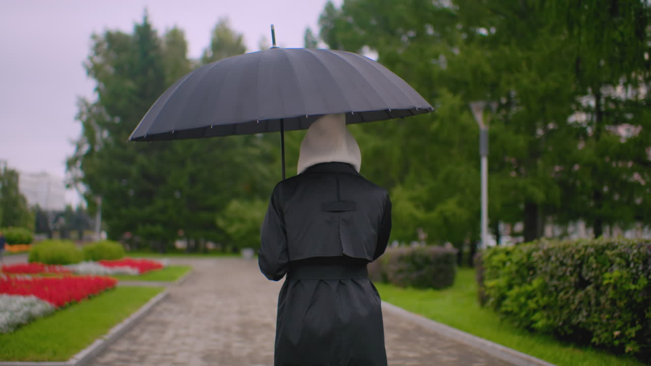 Back shot of woman in black coat with hood holds umbrella while walking on wet park path during rainy day, surrounded by blooming red flowers, lush greenery and benches creating calm serene