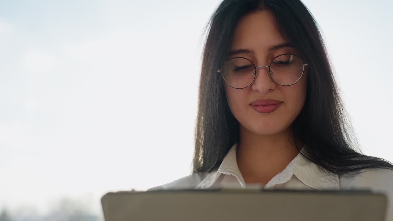 Close up woman smiling while using tablet with sunlight glow illuminating hair and face, blurred urban background, gentle lens flare capturing joyful moment in bright outdoor scene