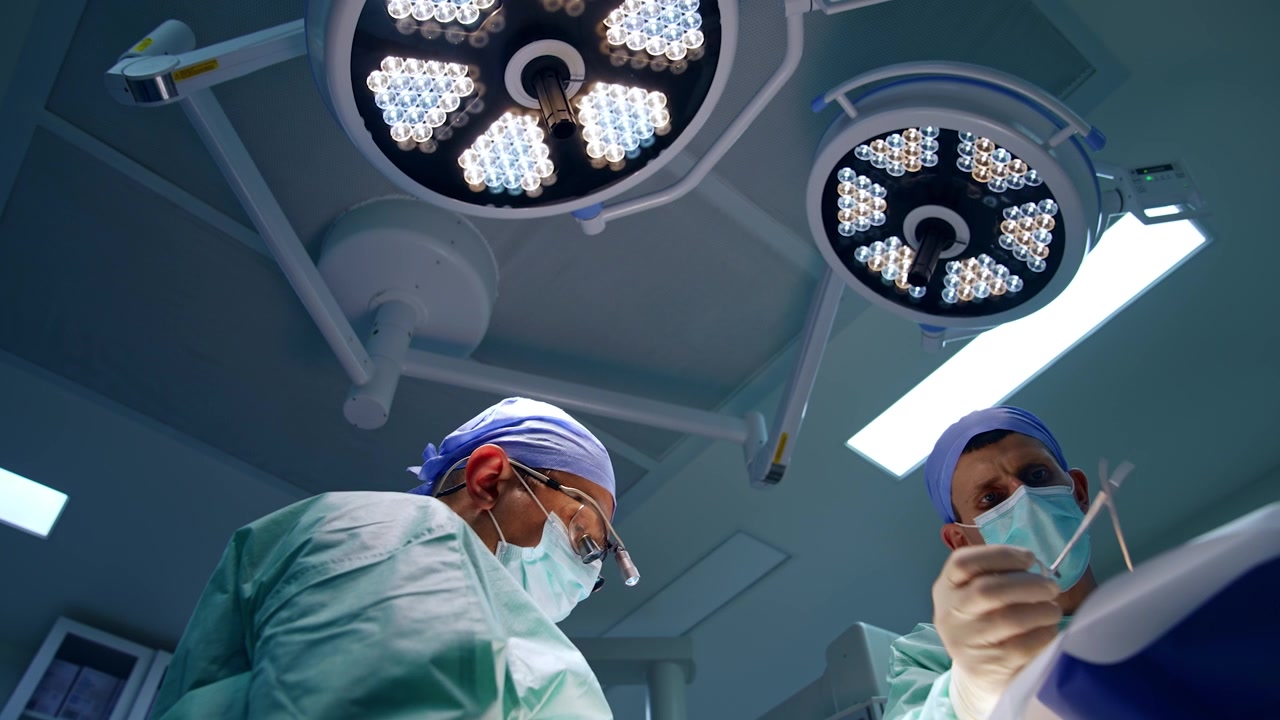Male Caucasian doctors in masks and caps work at operation. Low angle view on the medics operating the patient under the round lamps.
