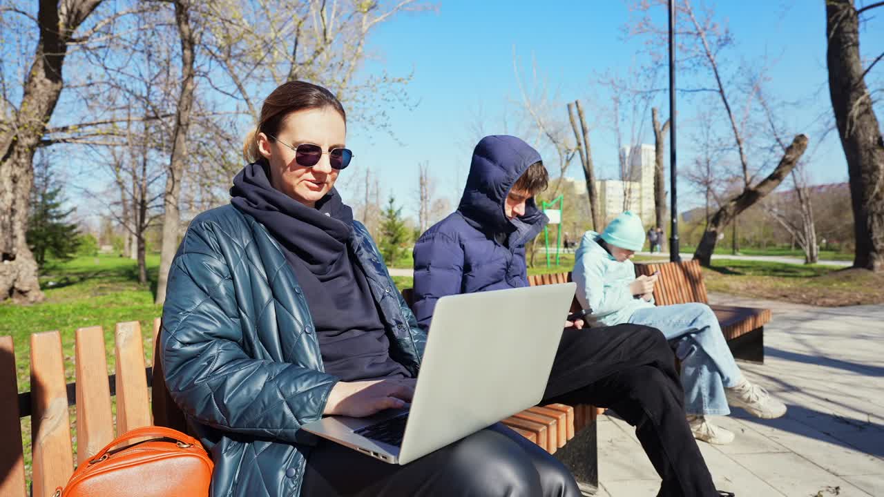 Kvinde arbejder på bærbar computer i parken med børn, der bruger telefoner