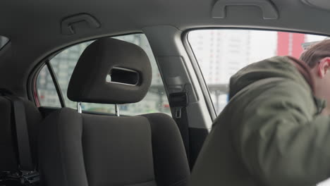teenager in green winter jacket opens car door, steps inside, and adjusts front seat forward, snowy urban scene visible outside vehicle through rear and side windows, modern city background