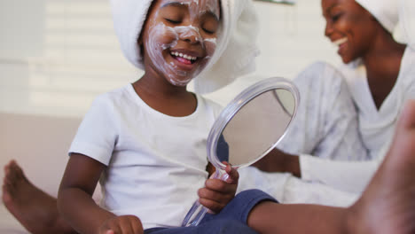 happy african american mother and daughter sitting on bed daughter putting facial cream