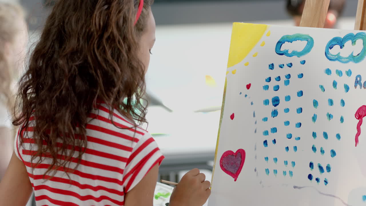 In a classroom at school, a biracial girl with curly brown hair is painting on a canvas